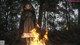 A woman standing in front of a fire in the woods.